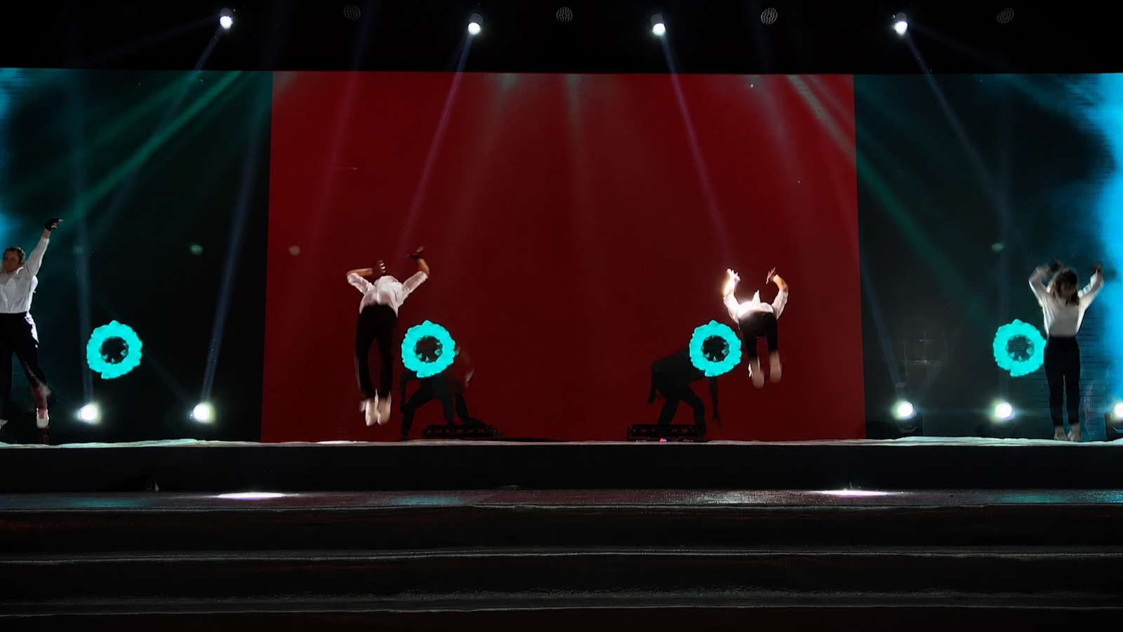 Dancers leaping across a stage in front of a deep red backdrop, teal beams crossing above them.