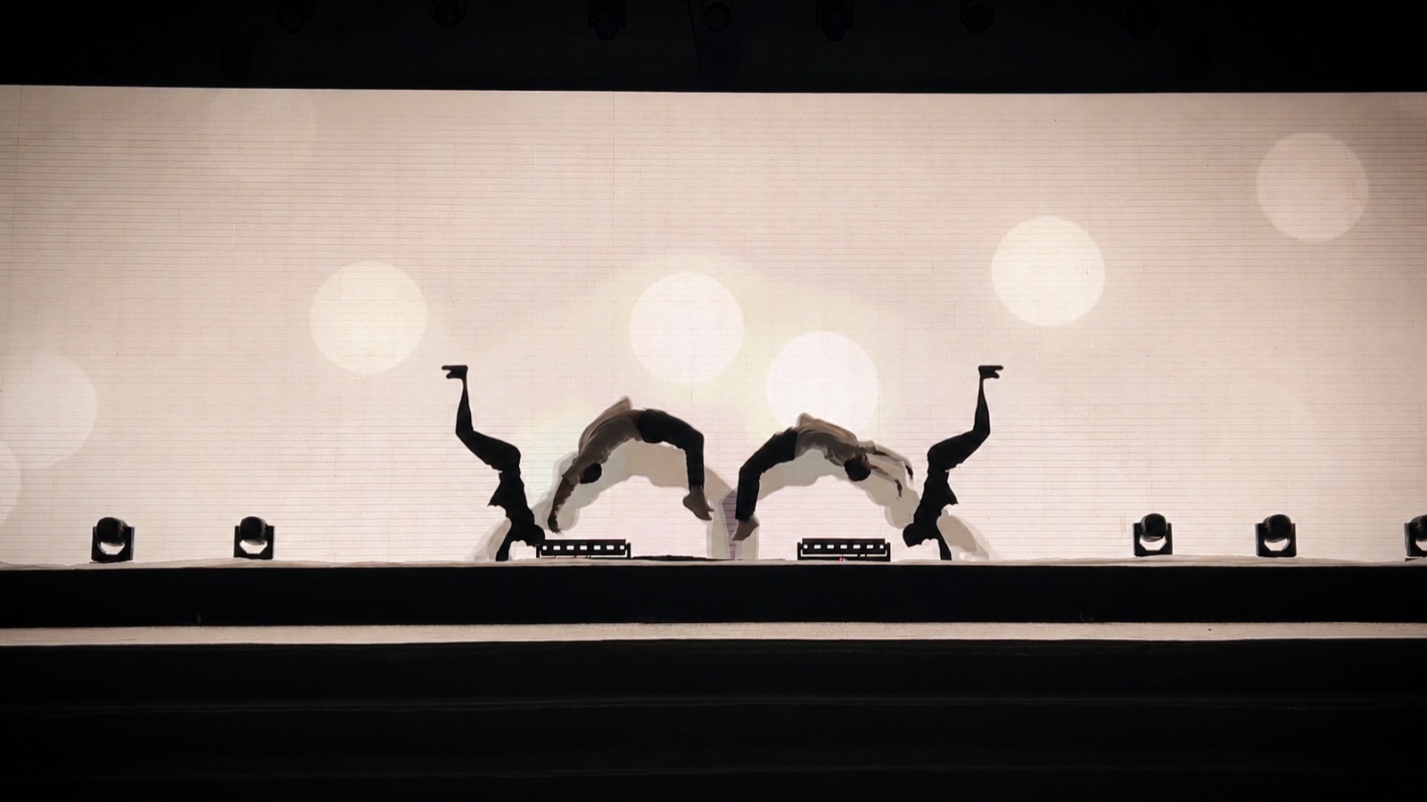Two performers in matching handstands, silhouetted against a pale screen scattered with soft light circles.