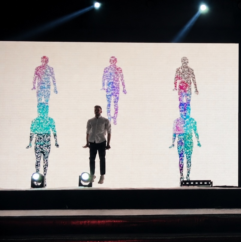A presenter walking downstage while the screen behind shows three human figures formed from particles.