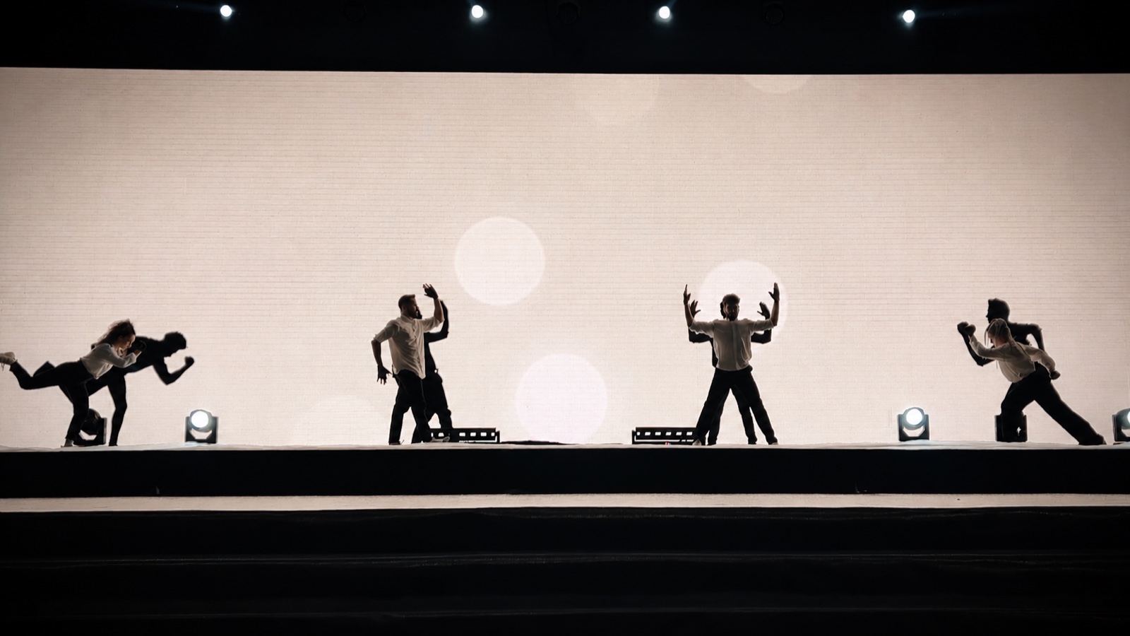 Four dancers spaced across a wide stage, silhouetted against a pale screen.
