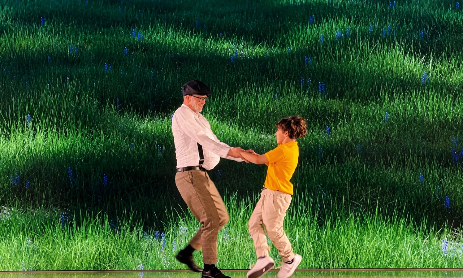An older performer and a young performer dancing together in front of a screen filled with tall green grass.