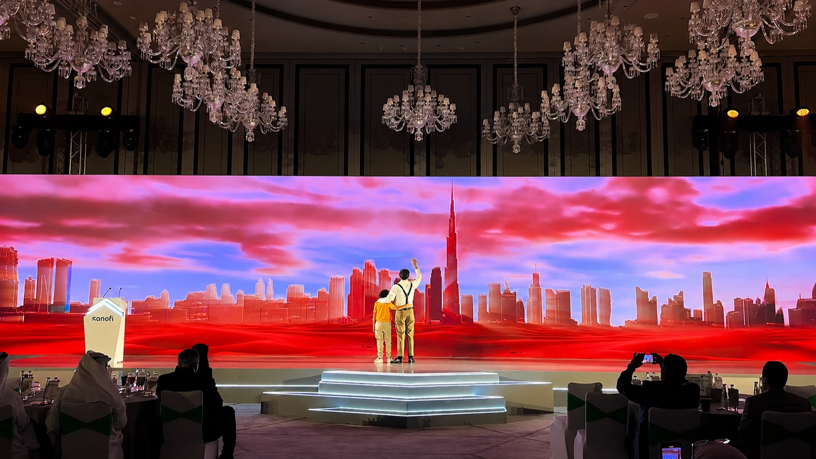 A ballroom stage under chandeliers: two performers on a low platform in front of a panoramic screen showing a red-sky Dubai skyline.