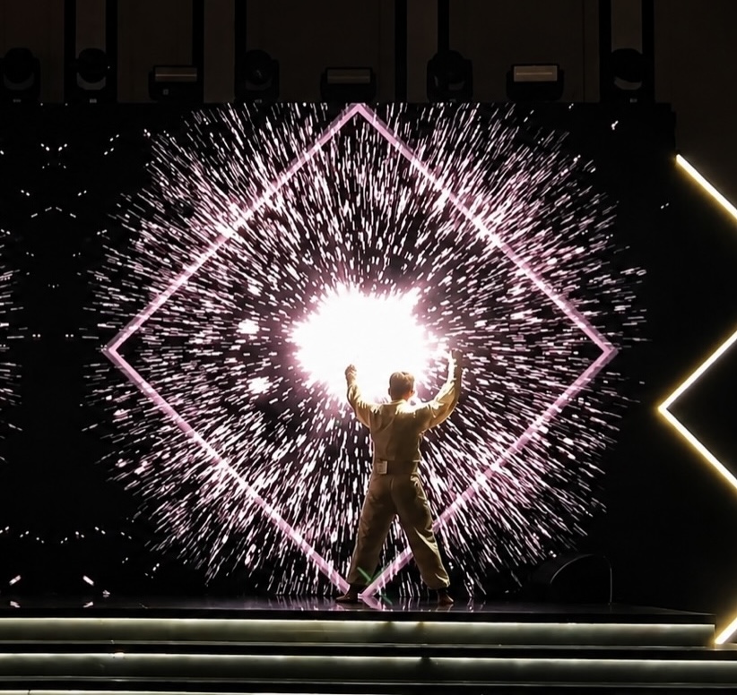 A performer silhouetted against a screen showing a bright burst inside a pink diamond outline.