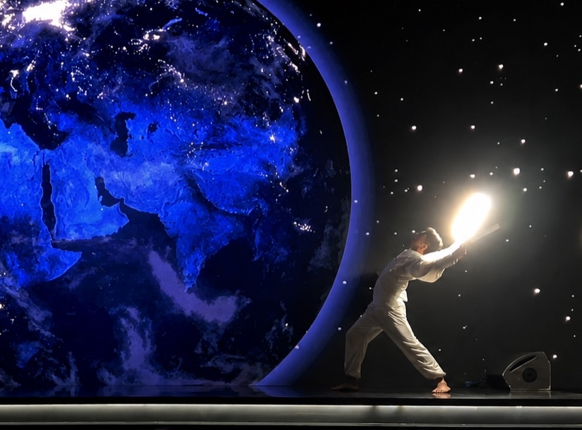 A performer reaching toward a point of light beside a screen showing the Earth from space.
