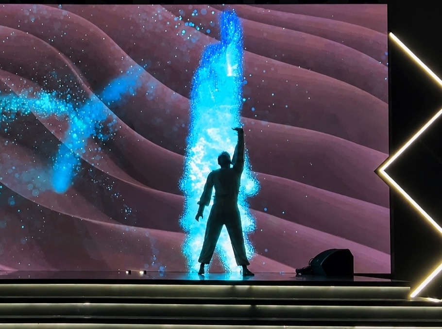 A performer raising one arm as a column of blue particles rises on the screen behind.
