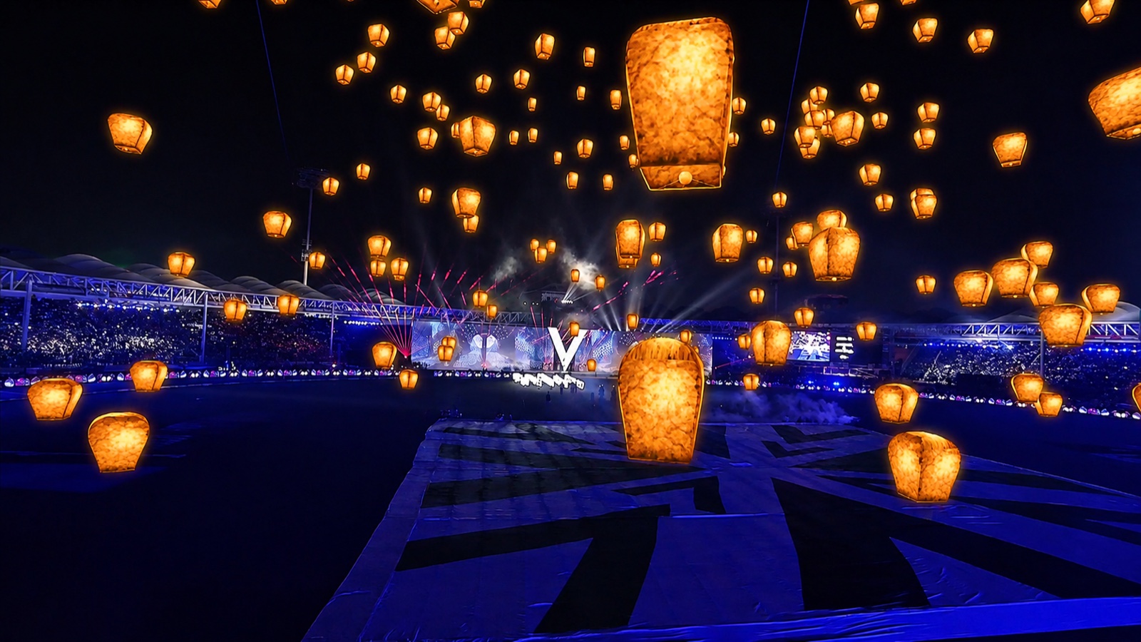 Hundreds of glowing lanterns rising above a floodlit stadium at night.