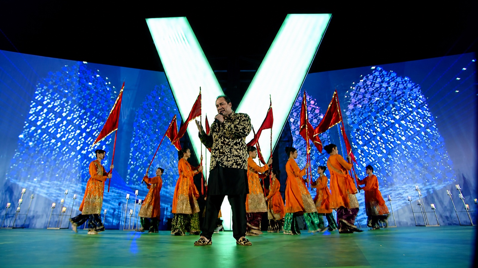 A singer performing on a stadium stage in front of a large illuminated V, flag-bearers in orange behind.