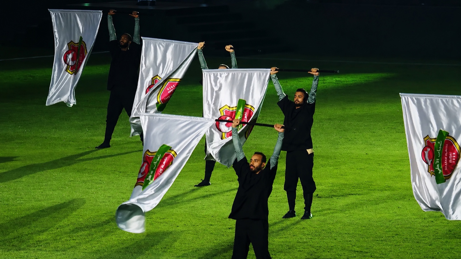 Performers in black spinning large white flags across a floodlit pitch.