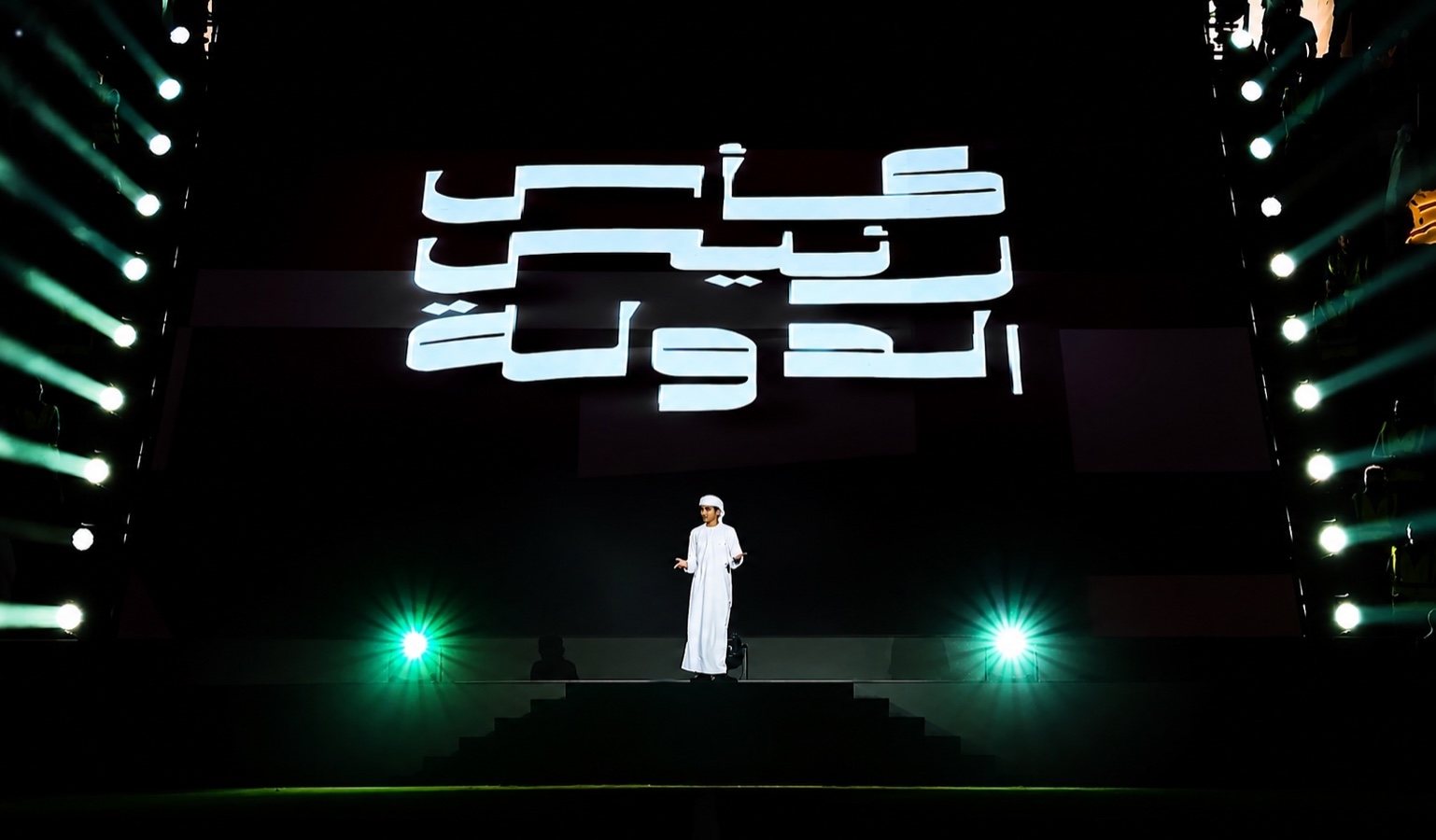 A performer in a white robe and headdress standing alone on a stage before large Arabic lettering.
