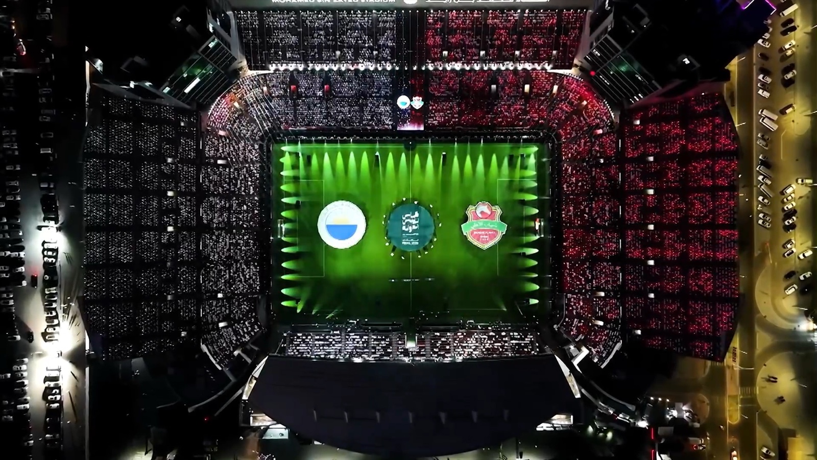 Aerial view of a floodlit stadium, three crest graphics laid across the green pitch.
