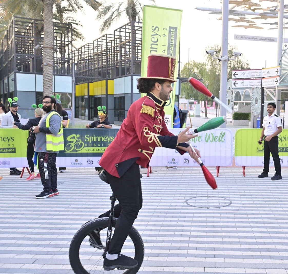 A performer in a red tailcoat and top hat juggling clubs while riding a unicycle among event visitors.