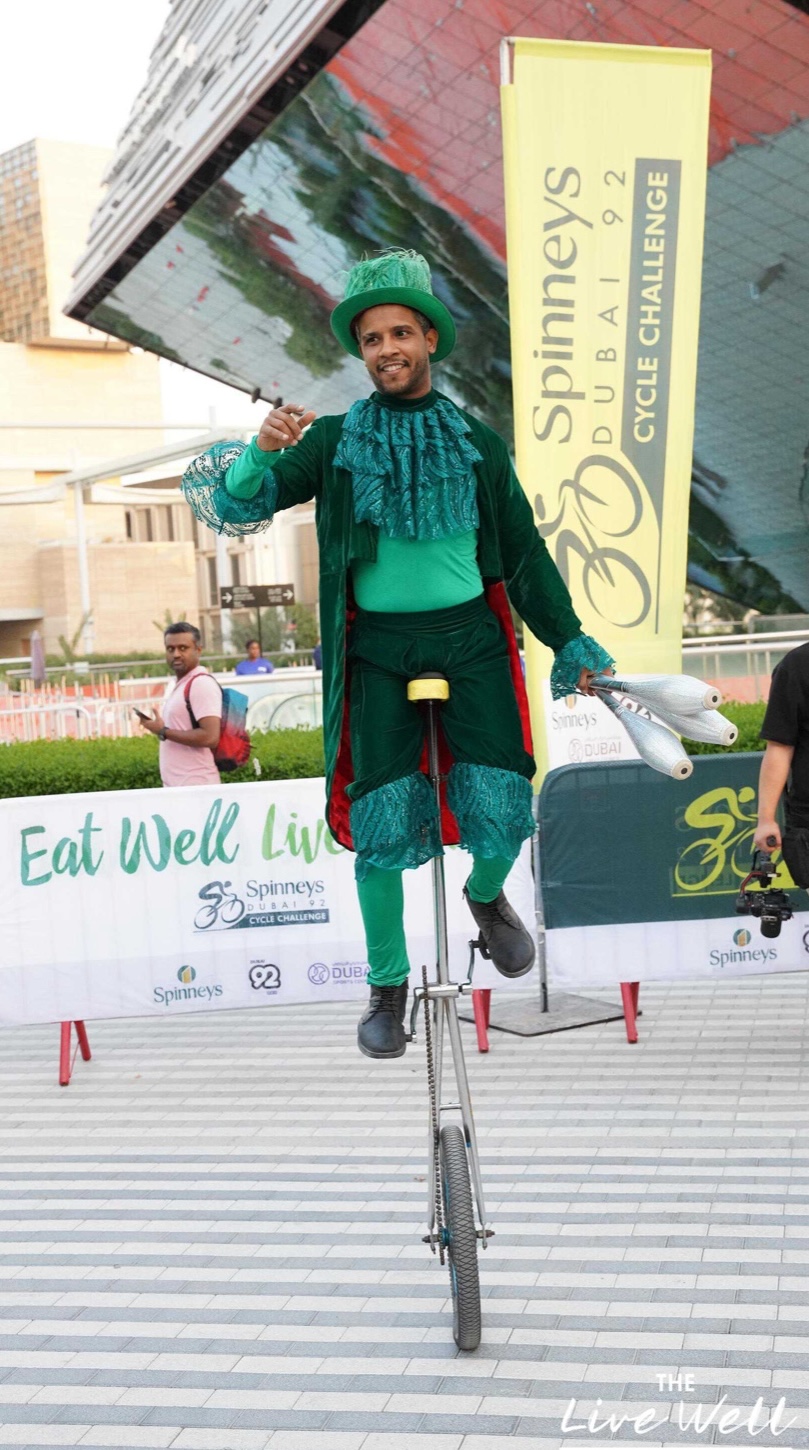 A performer in a green costume balanced on a tall unicycle beside event branding.