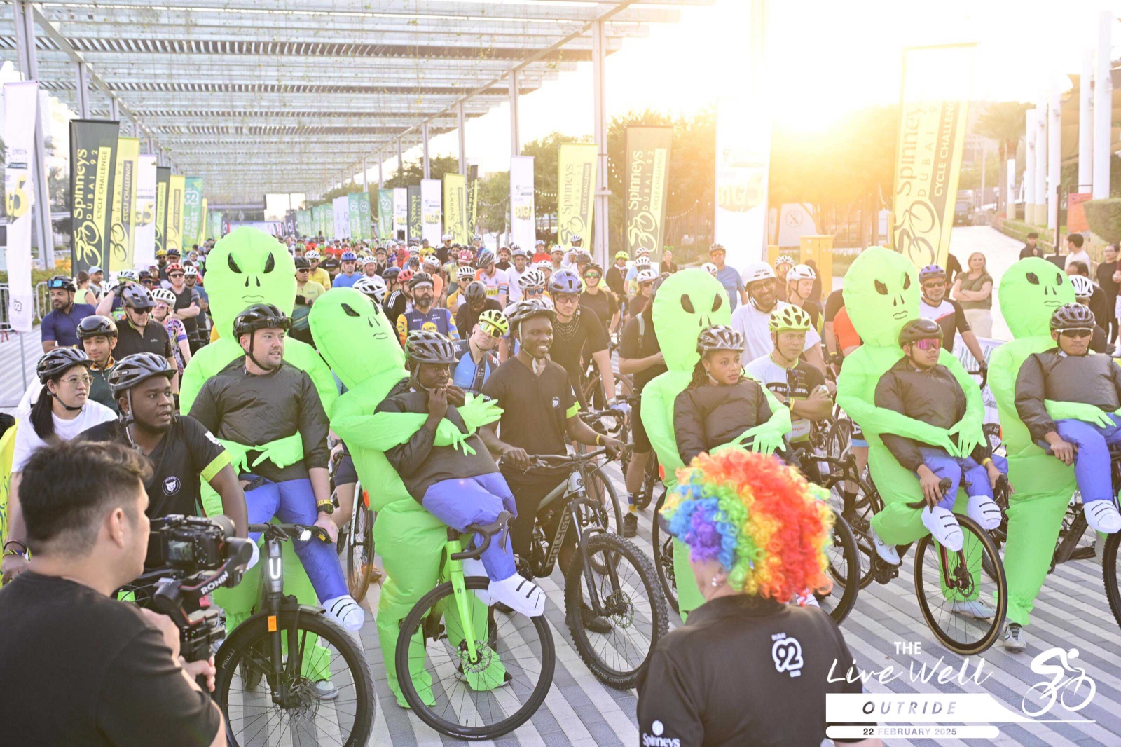 A mass ride setting off, several riders wearing green inflatable costumes among the field.